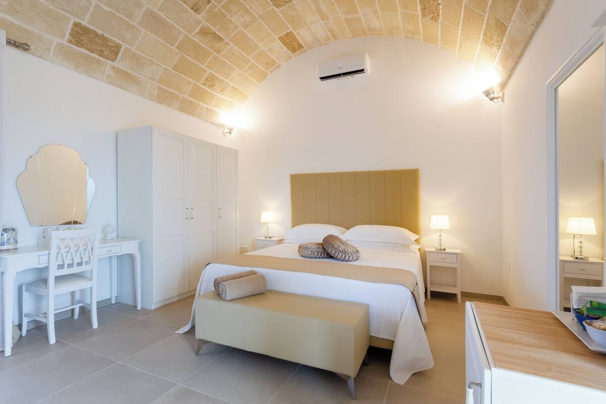 A hotel room with a bed, bedside tables, a desk, a chair, a bench, and a wardrobe.