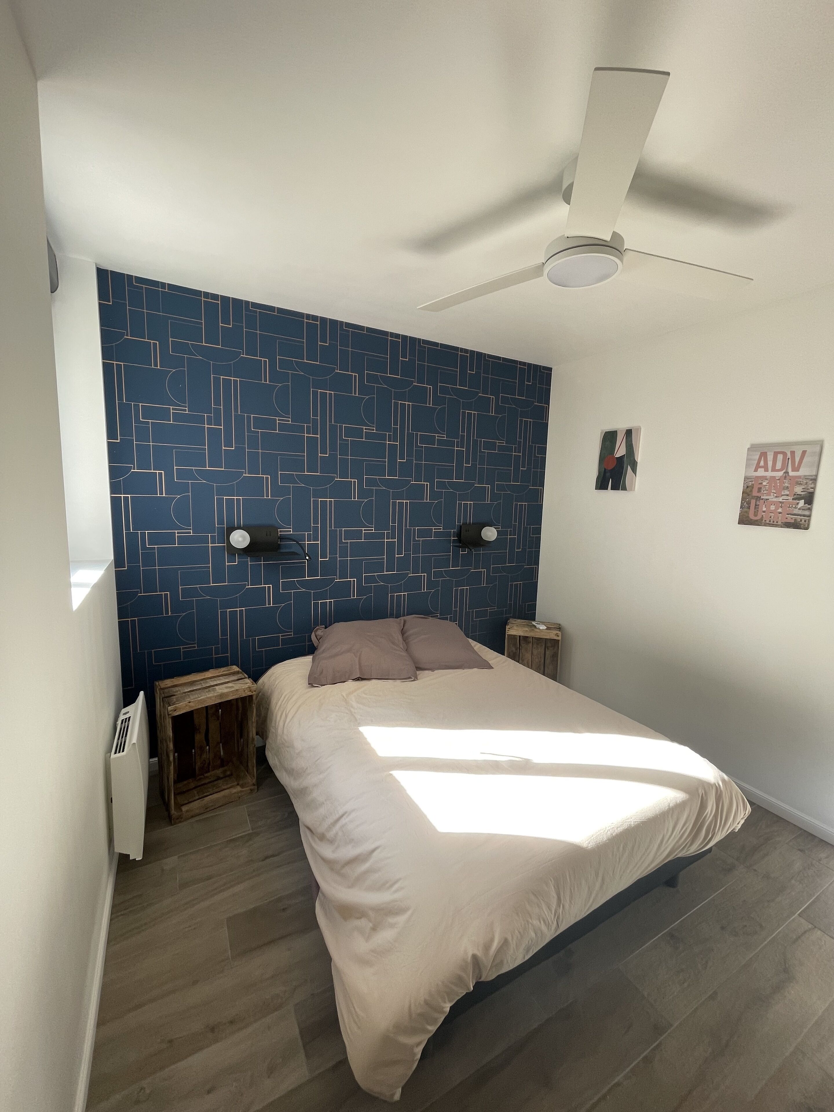 Une chambre à coucher avec un lit, un ventilateur de plafond, un mur orné d’un motif géométrique et deux petites tables de chevet en bois.