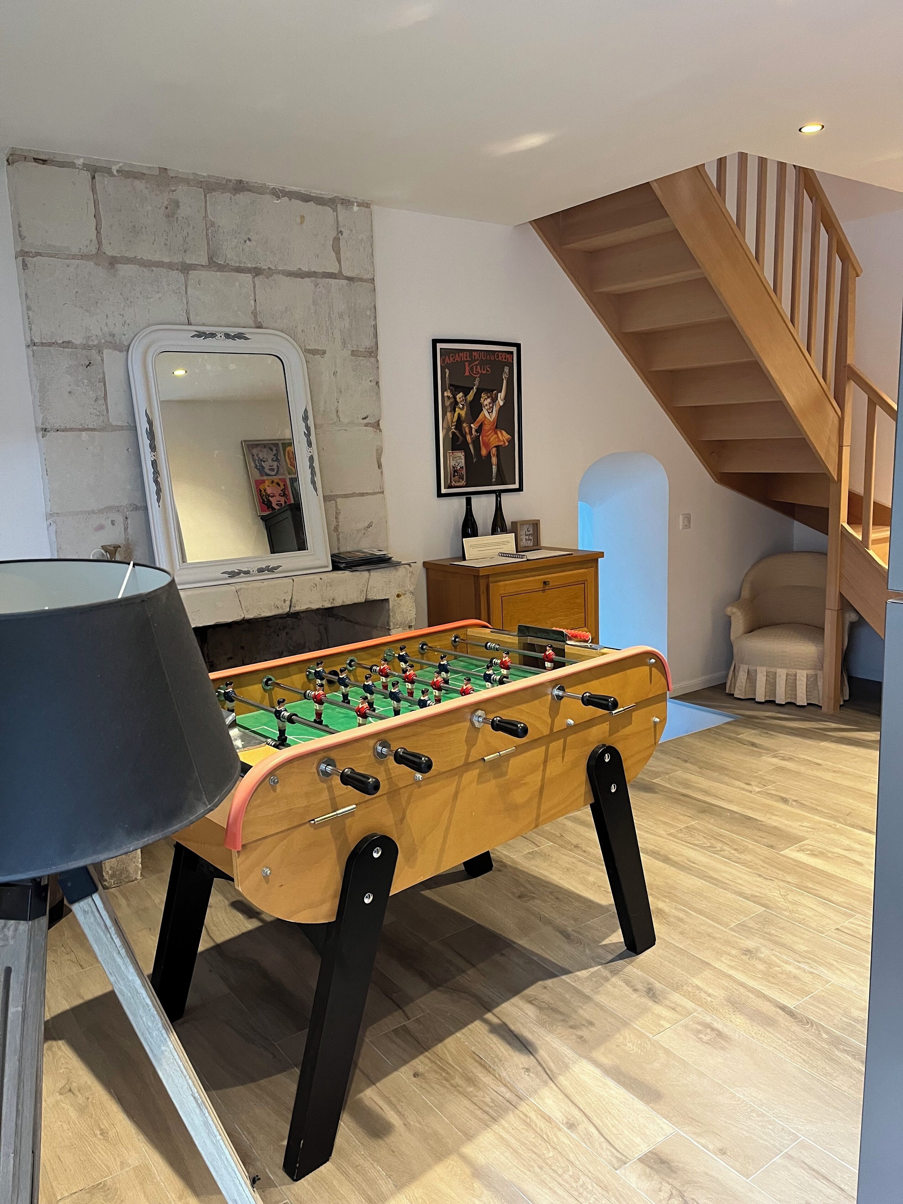 A room with a foosball table, a mirror, a framed picture, a wooden staircase, and a chair.