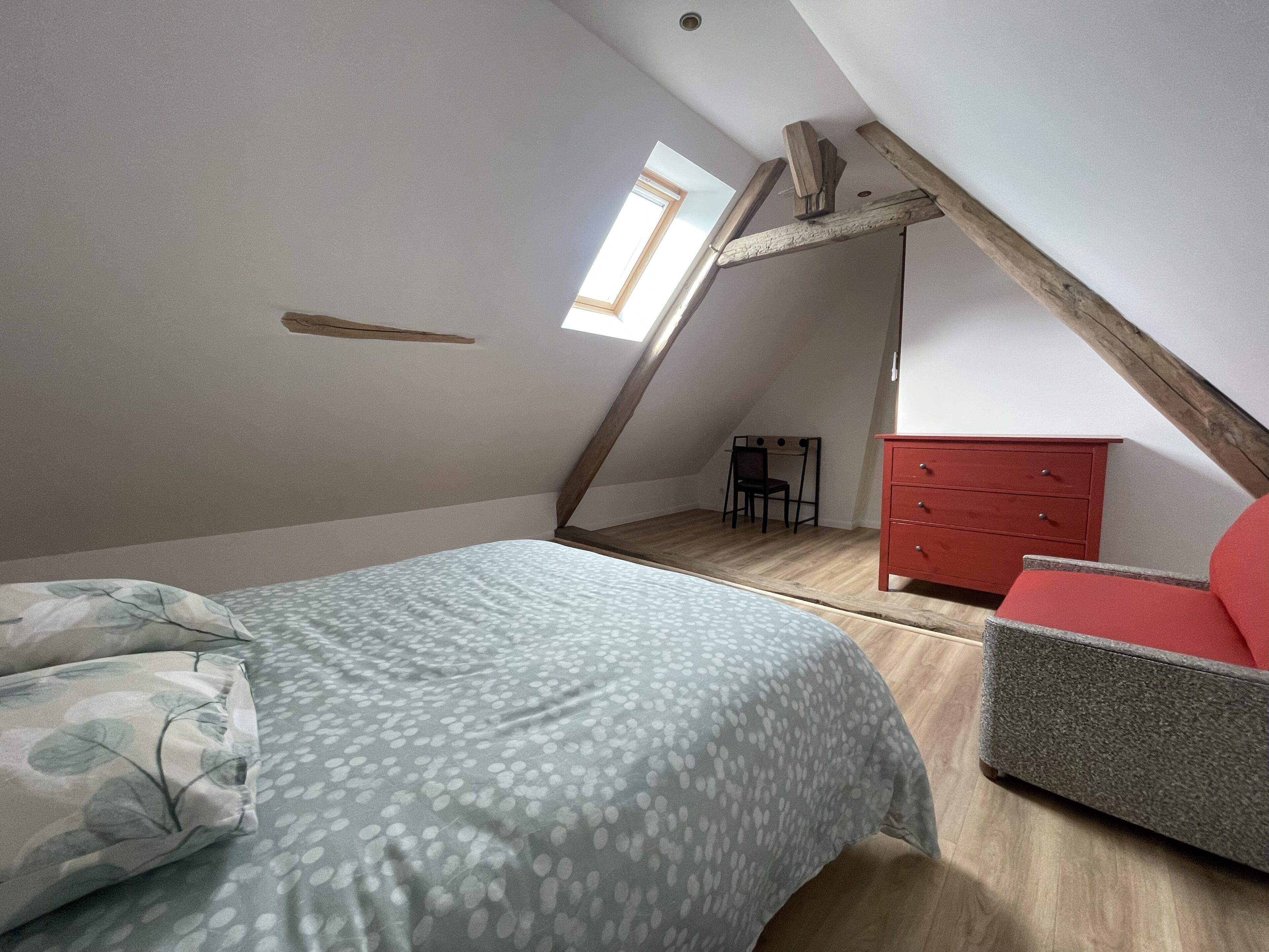 Quarto com teto inclinado, cama com roupa de cama estampada, cômoda vermelha e uma cadeira.