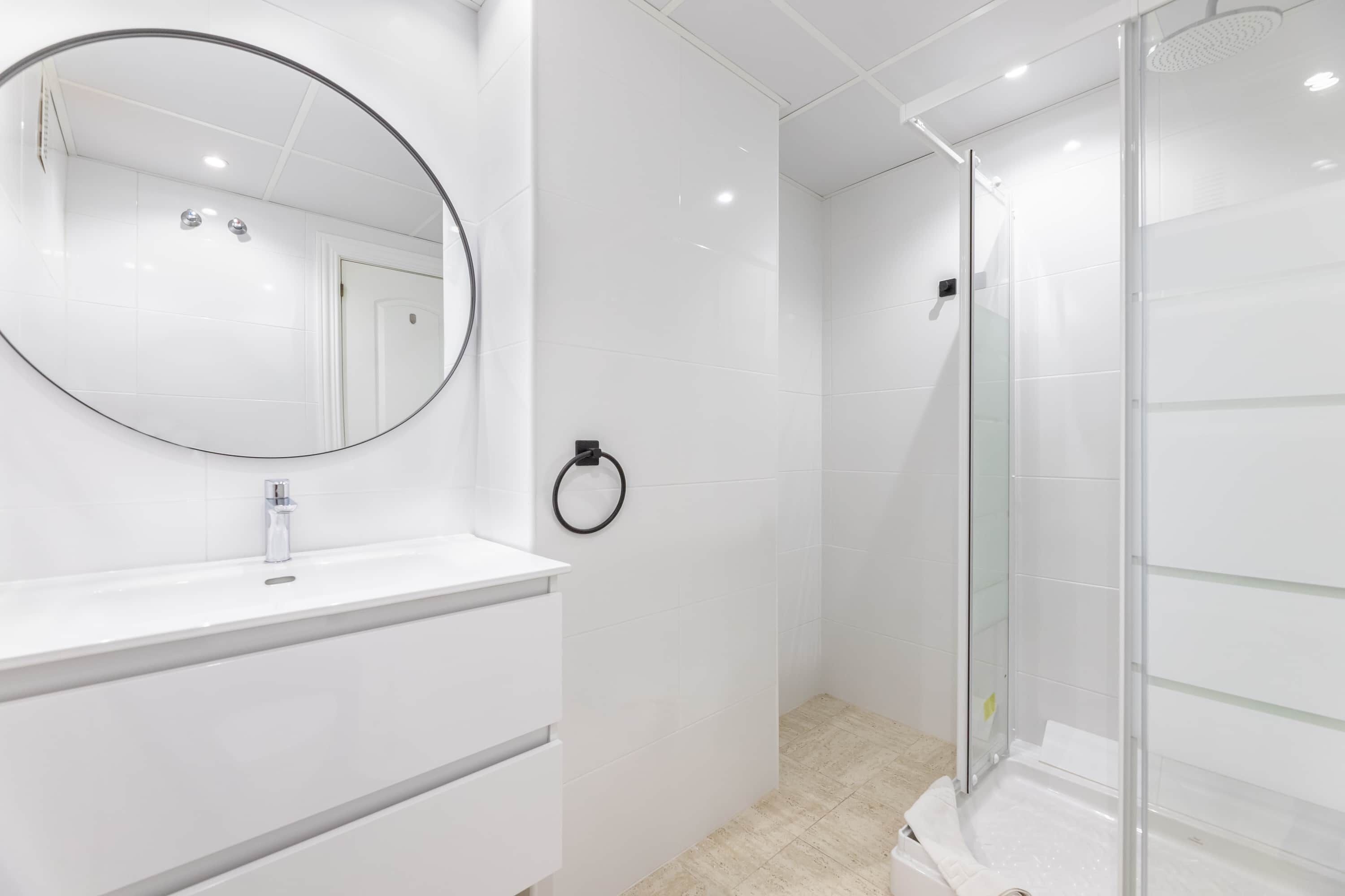 Un bany modern amb un moble de tocador blanc, un mirall rodó i una dutxa amb envàs de vidre.