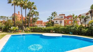 Ein Swimmingpool mit klarem, blauem Wasser, umgeben von Palmen und einem Gebäude im Hintergrund mit rot-weißen Wänden.