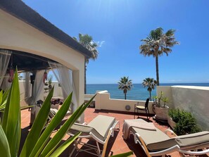 A terrace with lounge chairs, a table, and potted plants, overlooking a beach with palm trees and the ocean. - RDR318 - Spectacular views at beachfront townhouse (Estepona)