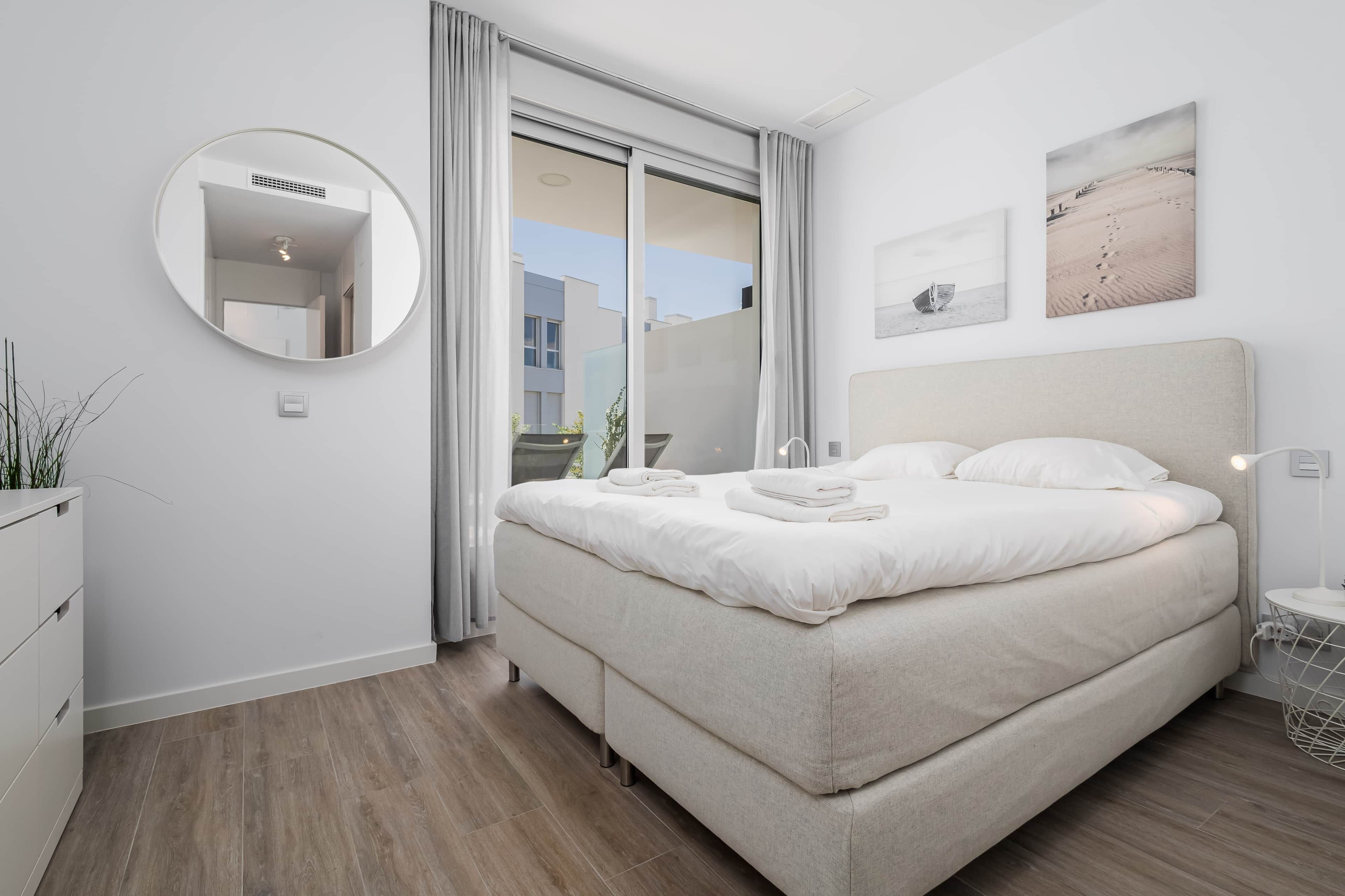 A modern bedroom with a large bed, a round mirror, and a view of the outside through a sliding door.