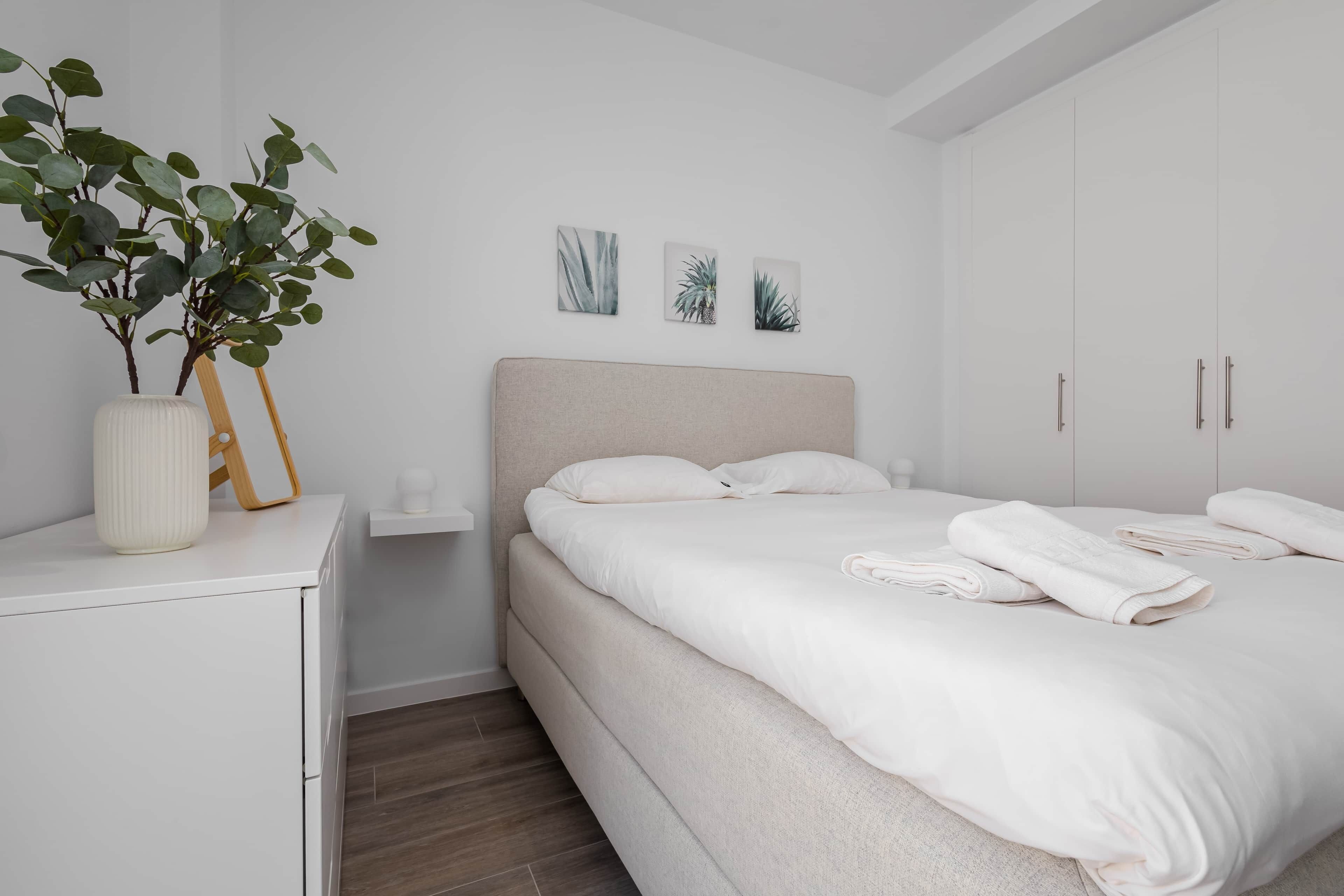 Een moderne slaapkamer met een bed, witte nachtkastjes, een kleine plant en wanddecoratie.