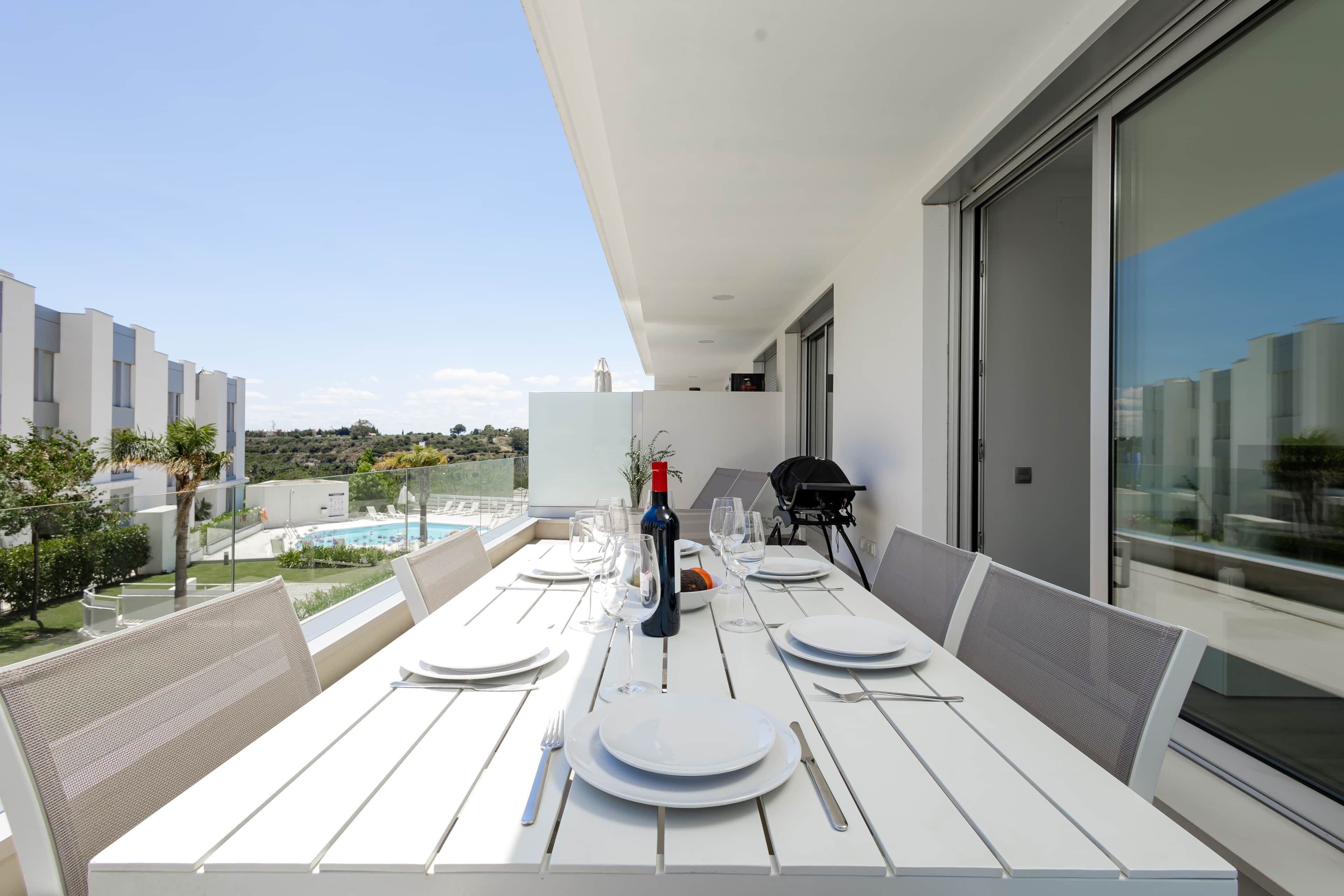 Ein Esstisch, gedeckt für eine Mahlzeit, befindet sich auf einem Balkon mit Blick auf einen Swimmingpool und Gebäude.