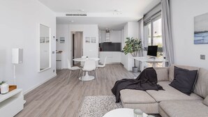 A modern, well-lit living room with a sofa, a coffee table, and a dining area with a table and chairs. There is a kitchen in the background with a large window. - RDR317 - Stylish 3 bedroom Le Mirage close to beach (Estepona)