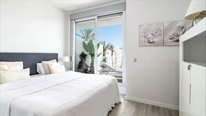 Une chambre avec un grand lit, une table de chevet, une lampe et une vue sur les palmiers et les immeubles, visible à travers une porte-fenêtre coulissante.