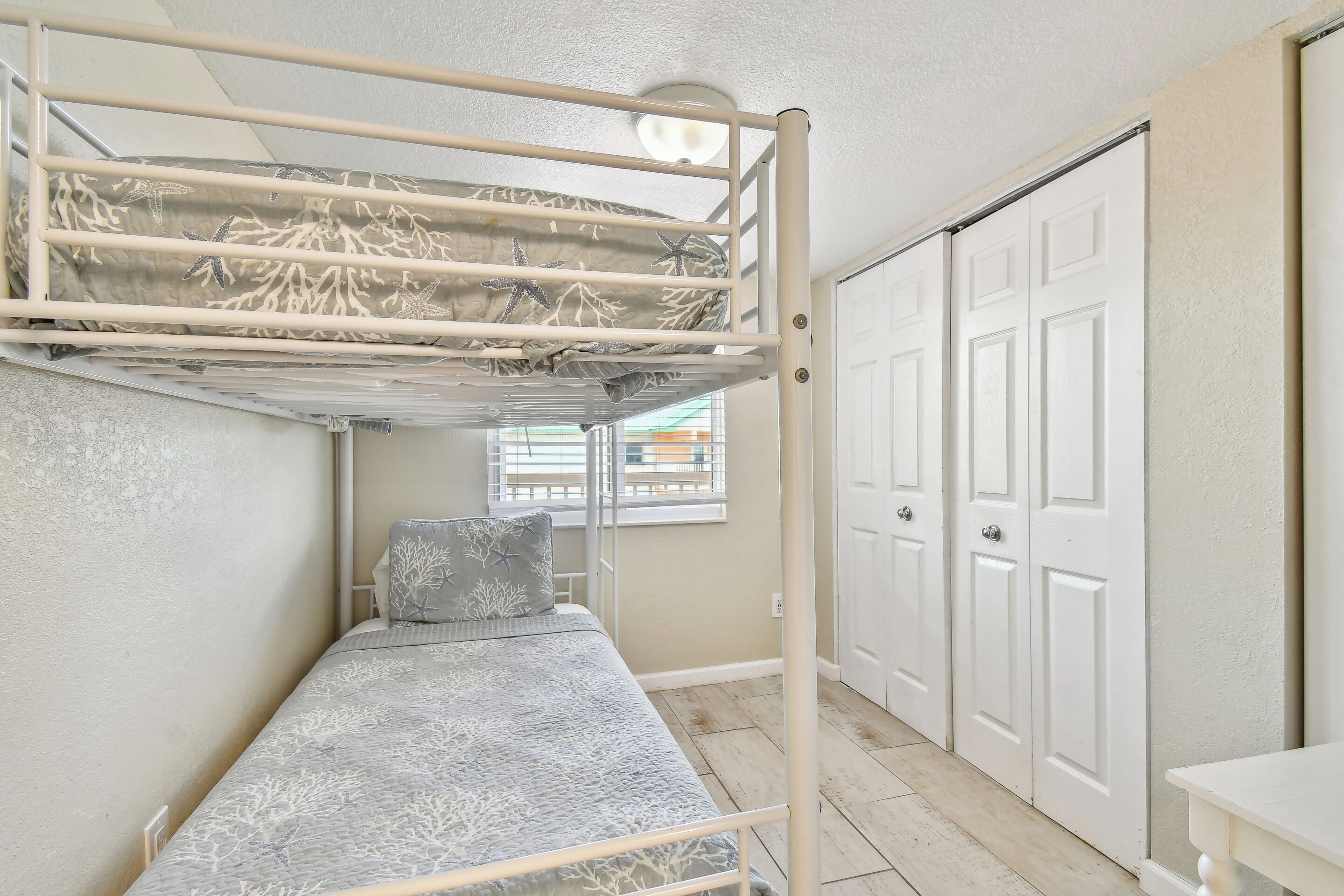 Un lit superposé avec une couvre-lit à motifs, un lit simple avec un oreiller, une porte blanche et une fenêtre avec des stores.