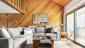 Un salón moderno con techo de madera, un sofá gris, una mesa de centro con cubierta de vidrio y un reposapiés estampado.