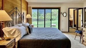 Un dormitorio con cama, lámparas de noche, cabecera de madera, vista a través de una puerta de cristal y una silla.