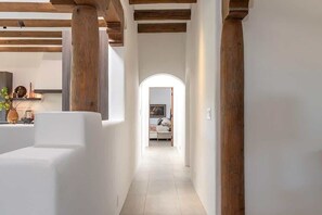 A hallway with wooden beams, white walls, and a tiled floor leading to a room with a bed and a window.