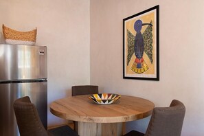 A kitchen with a stainless steel refrigerator, a woven basket, a round wooden table with a colorful bowl, and a framed artwork on the wall.