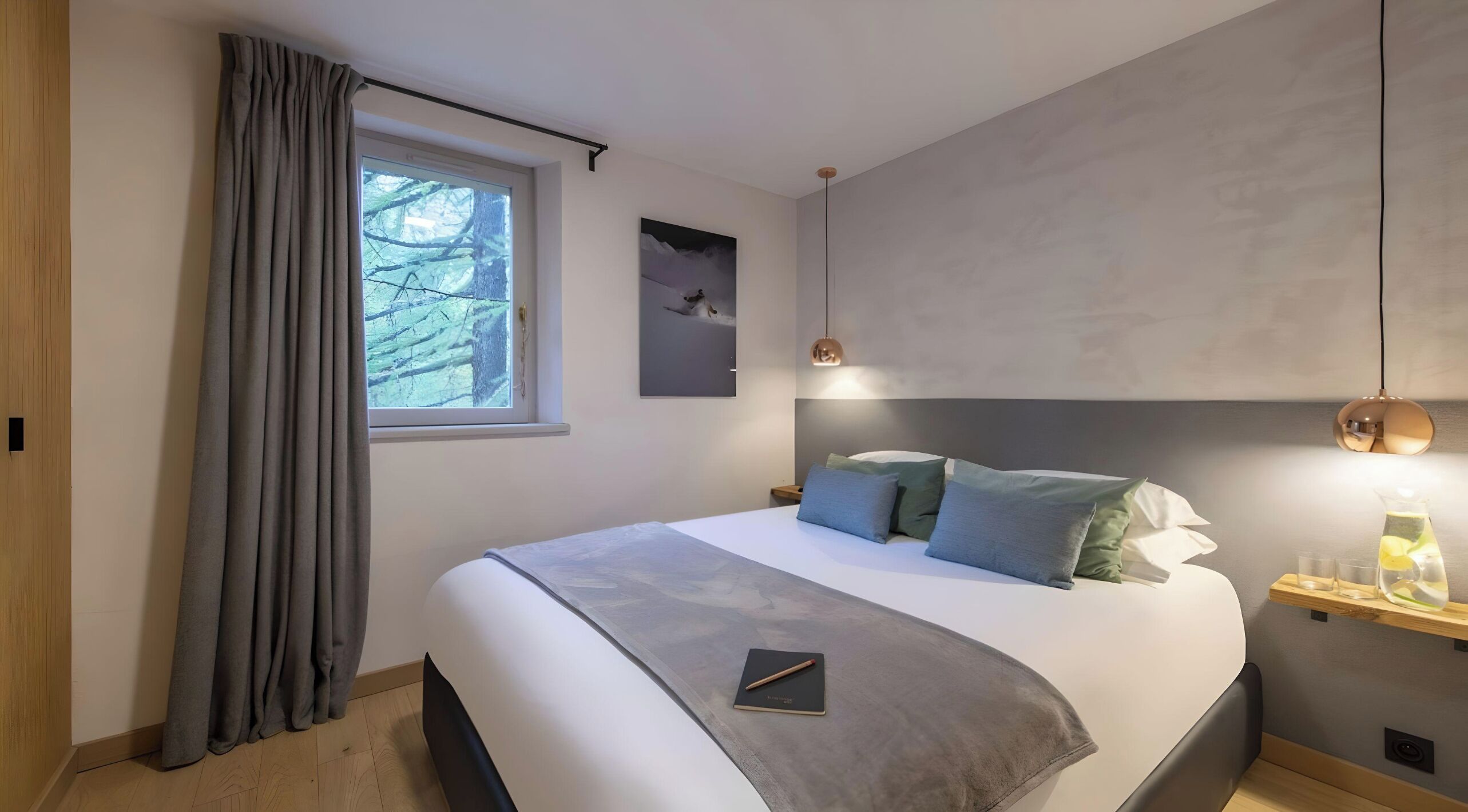 Een moderne slaapkamer met een groot bed, grijze gordijnen, een raam met uitzicht op bomen, een nachtkastje met een lamp en een klein plankje met glazen.