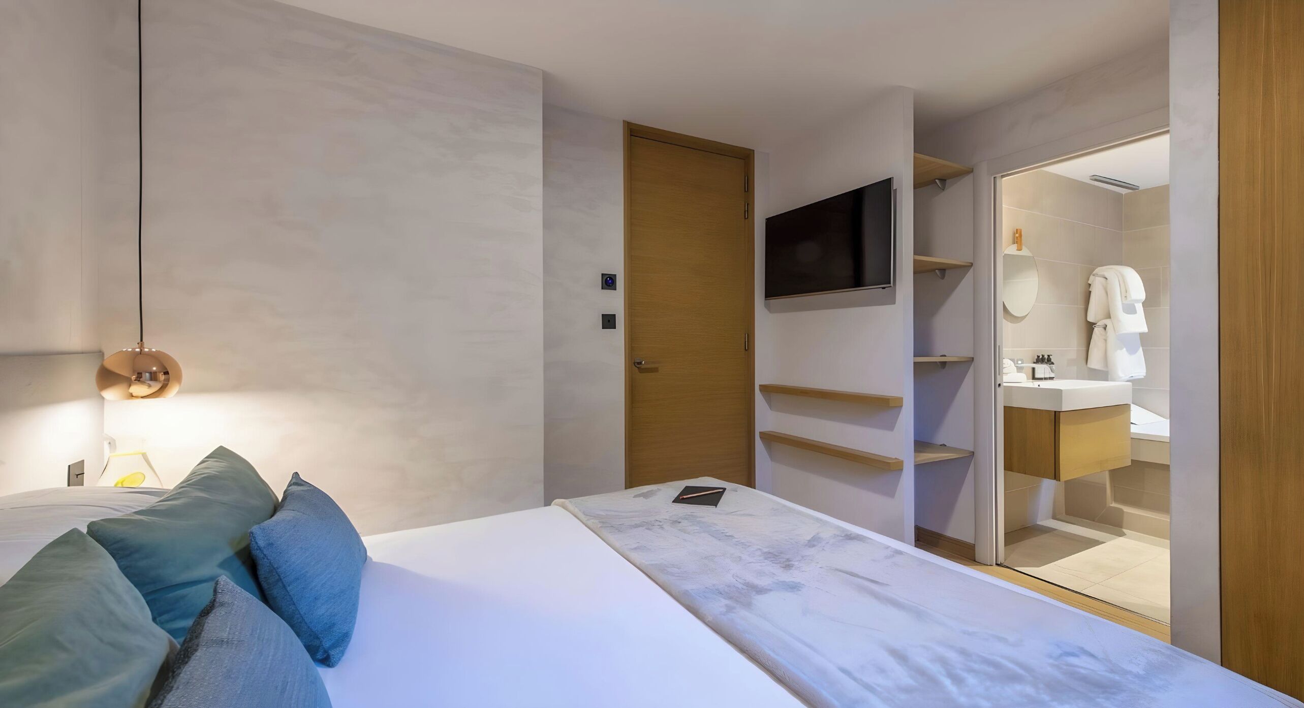 Une chambre à coucher moderne, avec un lit, un téléviseur fixé au mur et une salle de bain visible à travers une porte ouverte.