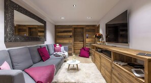 A modern living room with a grey sofa, wooden panel walls, a large TV, and a wooden console with electronic devices.