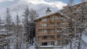 Un edifici de fusta de diversos pisos amb balcons, envoltat d'arbres coberts de neu i muntanyes al fons.
