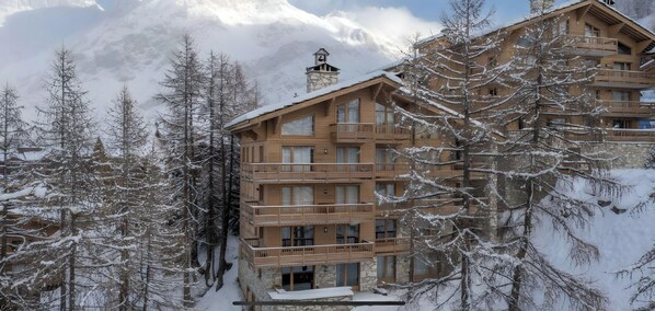 A multi-story wooden building with balconies, surrounded by snow-covered trees and mountains in the background.