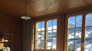 A wooden cabin room with a bed, a fur rug, and large windows offering a snowy mountain view. - Cozy family apartment with stove, gallery and magnificent alpine view (Adelboden)