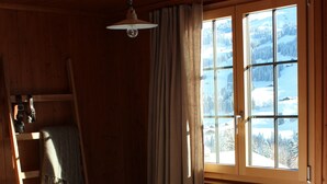 A cozy room with a bed, a window with a view of snow-covered mountains, a wooden ladder, and a hanging lamp. - Cozy family apartment with stove, gallery and magnificent alpine view (Adelboden)