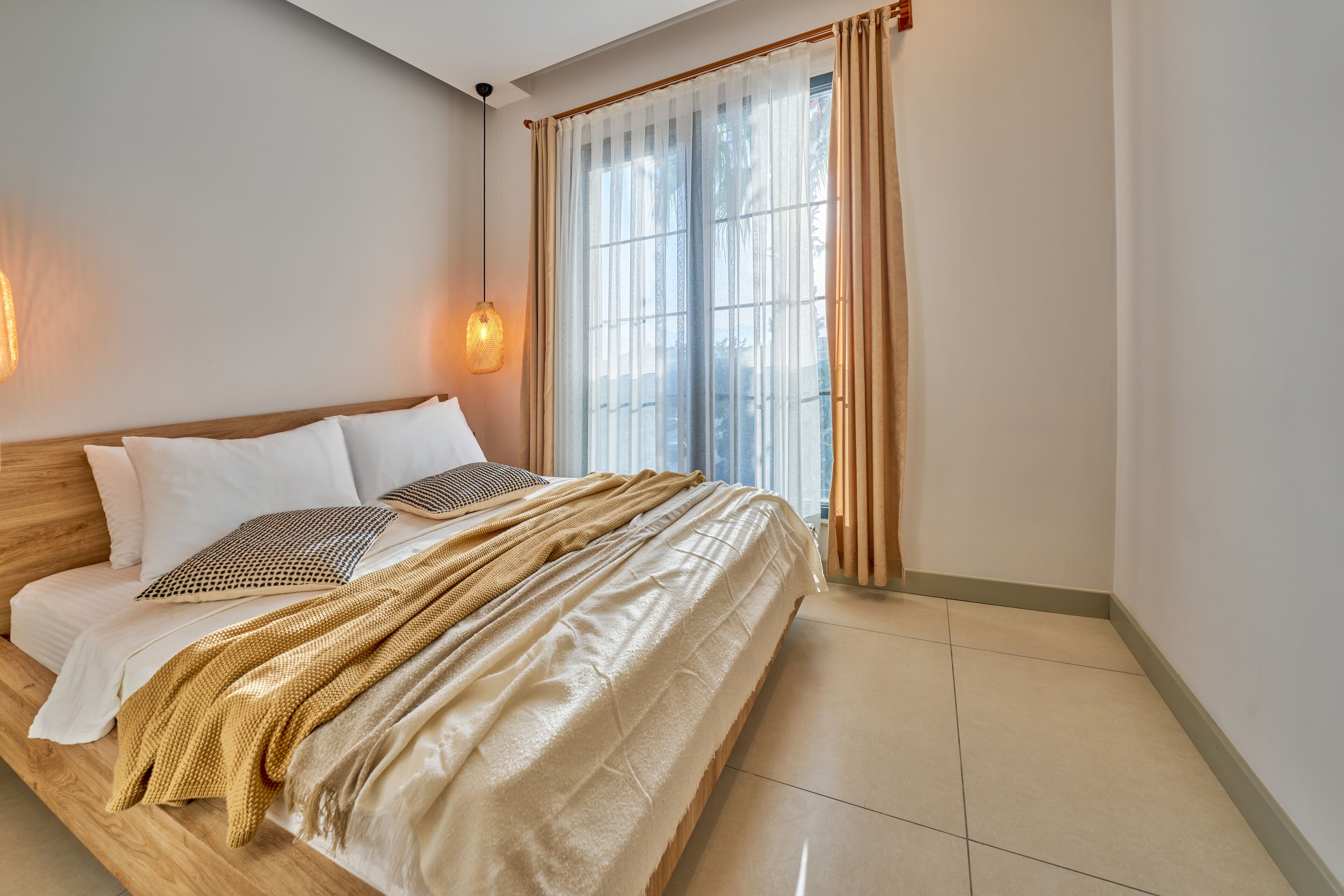 A neatly made bed with a wooden headboard, a large window with sheer curtains, and a hanging light fixture.