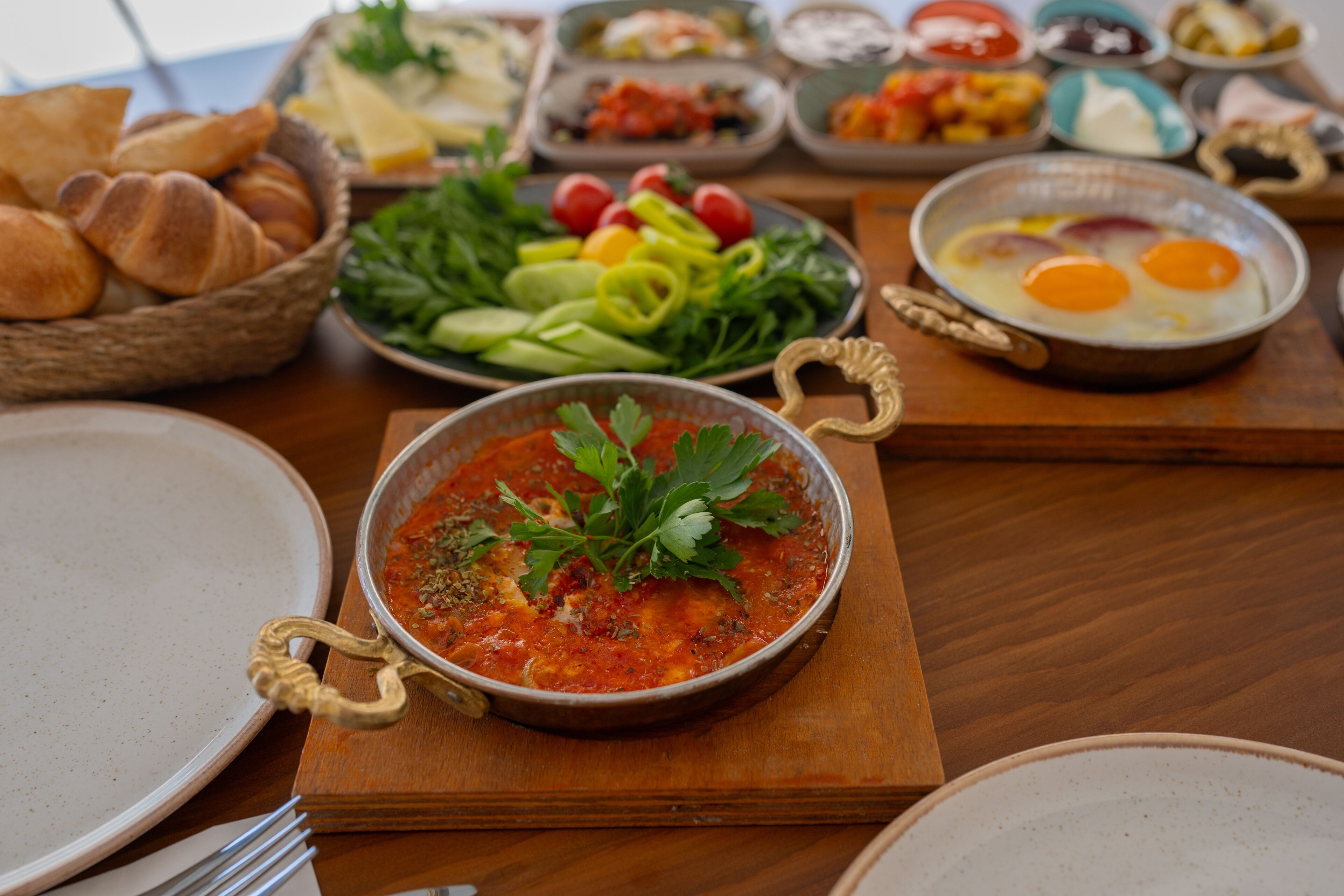 A dining table with various dishes including a bowl of tomato-based soup garnished with parsley, a plate of eggs, and a variety of vegetables and bread.