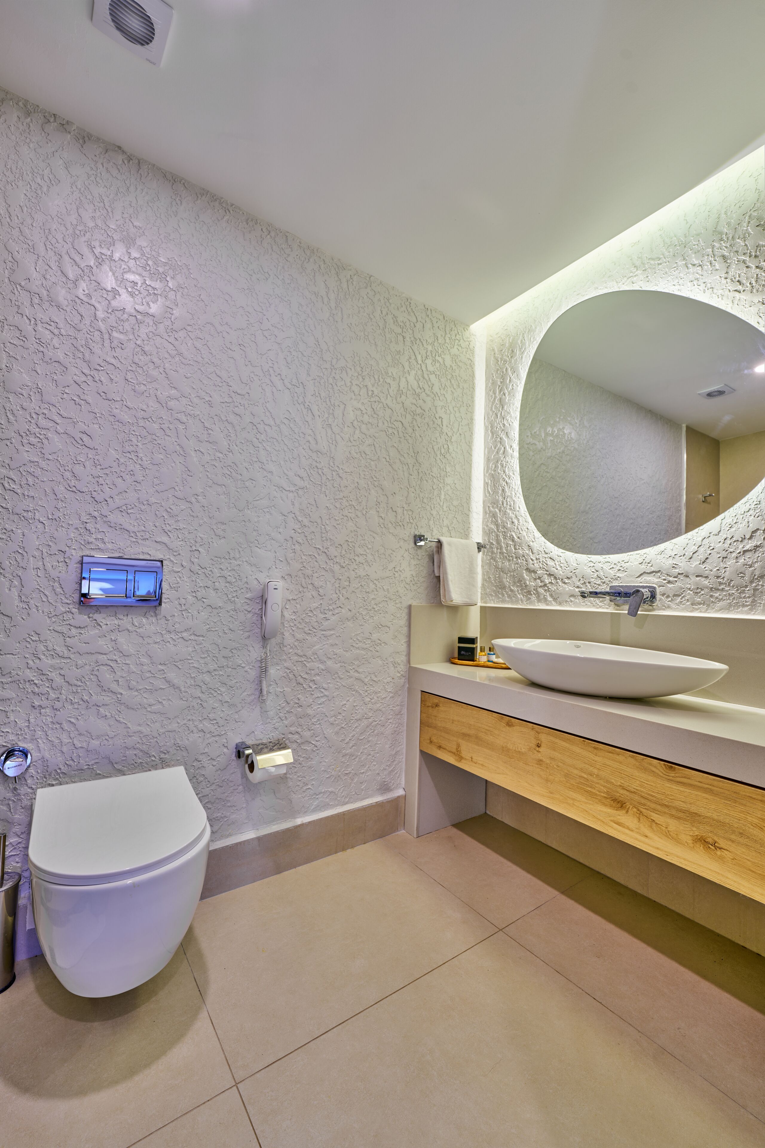A bathroom with a textured wall, a white toilet, a sink with a round mirror, and a wooden vanity.