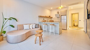 Una sala moderna con un sofá curvo, dos taburetes de mimbre y una zona de cocina con barra.