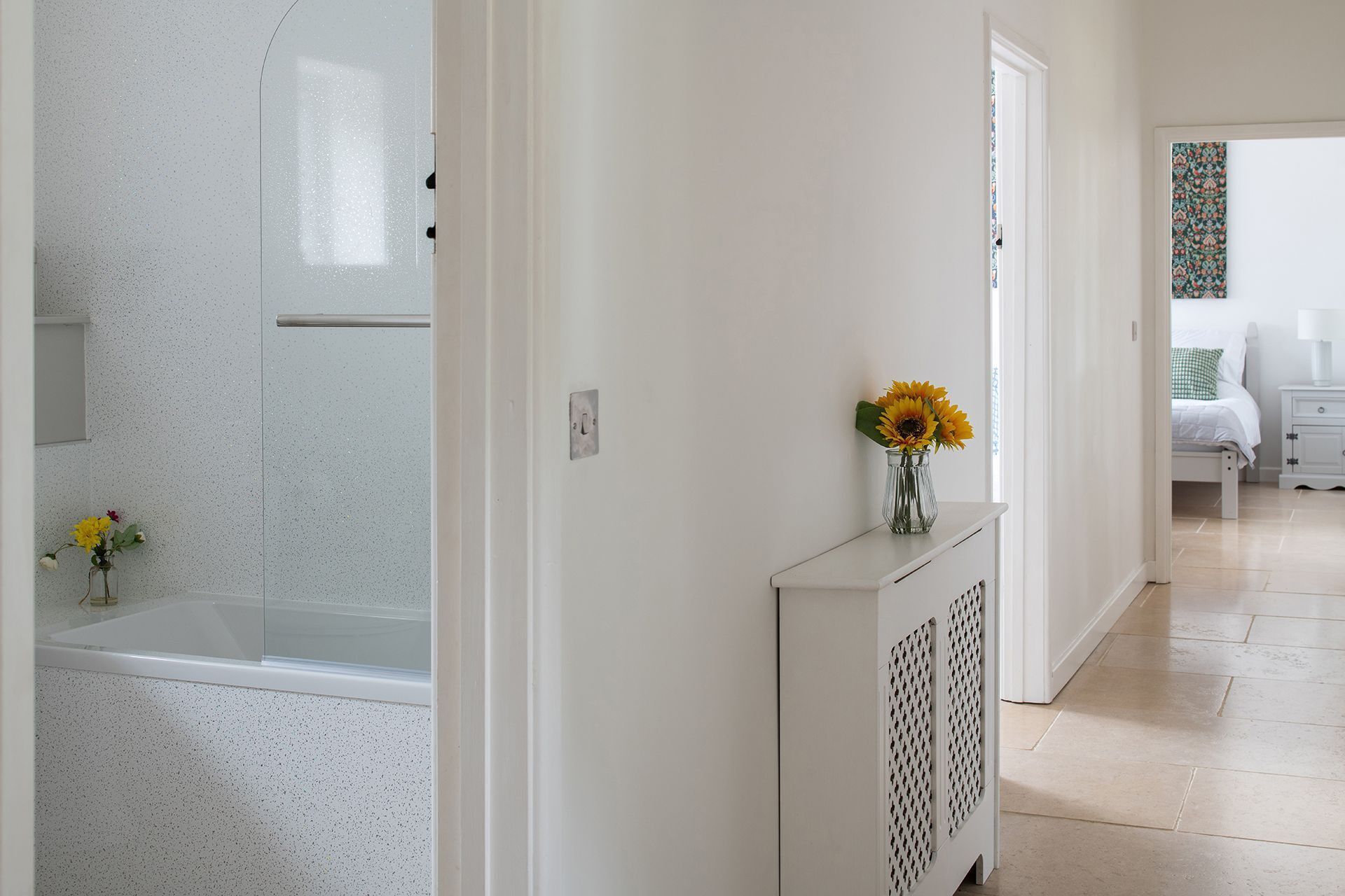 Um corredor com um radiador branco, um vaso com girassóis e um vislumbre de um quarto com cama e mesa de cabeceira.