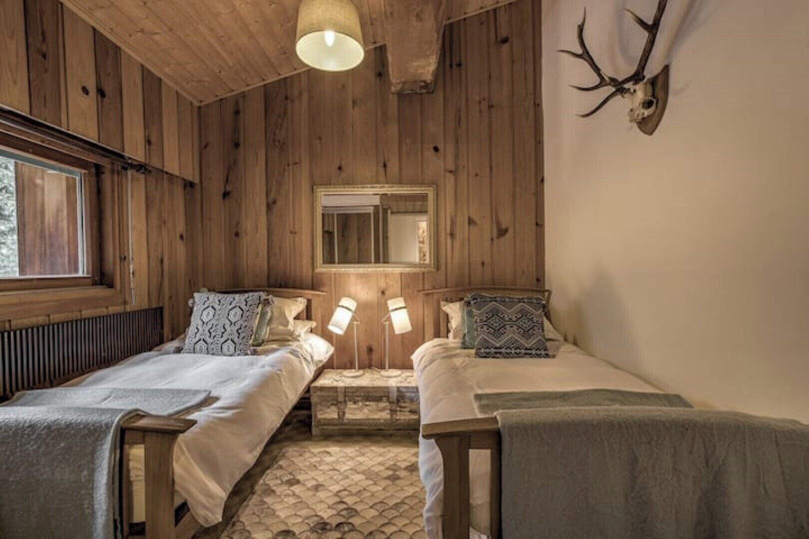 A wooden cabin room with two beds, a mirror, and a mounted deer skull.