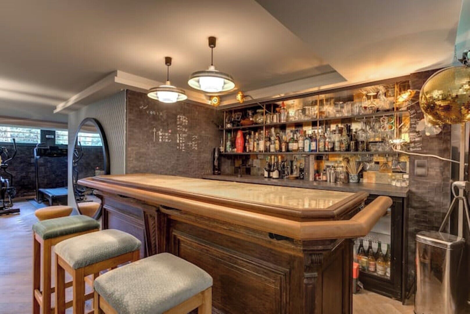 Een bar met een houten toog, barstoelen en een goed gevulde plank met flessen.