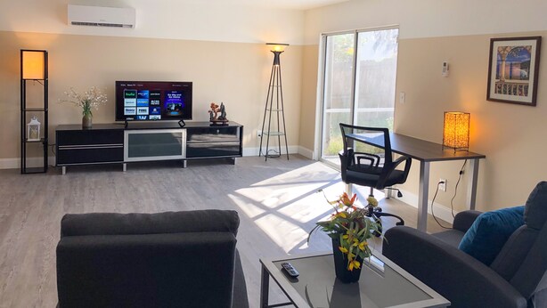 A modern living room with a flat-screen TV, a floor lamp, a dining table, and a sofa. - Lazy Cuckoo Inn (Fort Myers Beach)