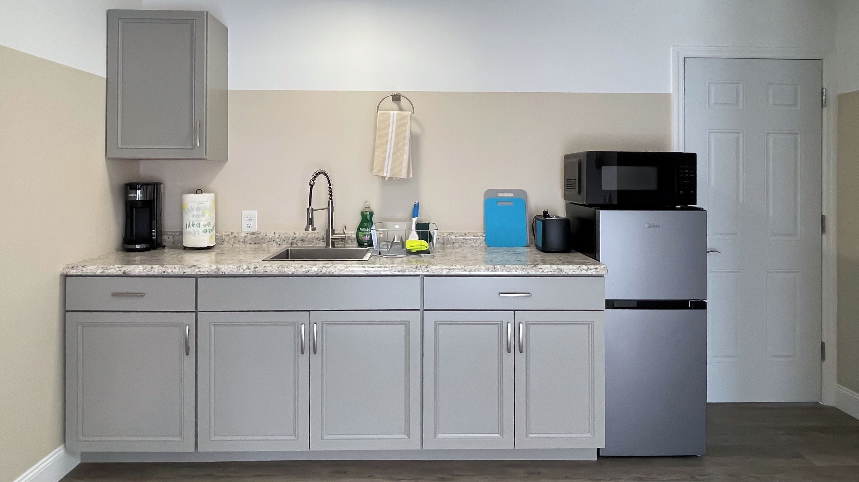 A kitchen with grey cabinets, a stainless steel sink, a coffee maker, a microwave, and a refrigerator.