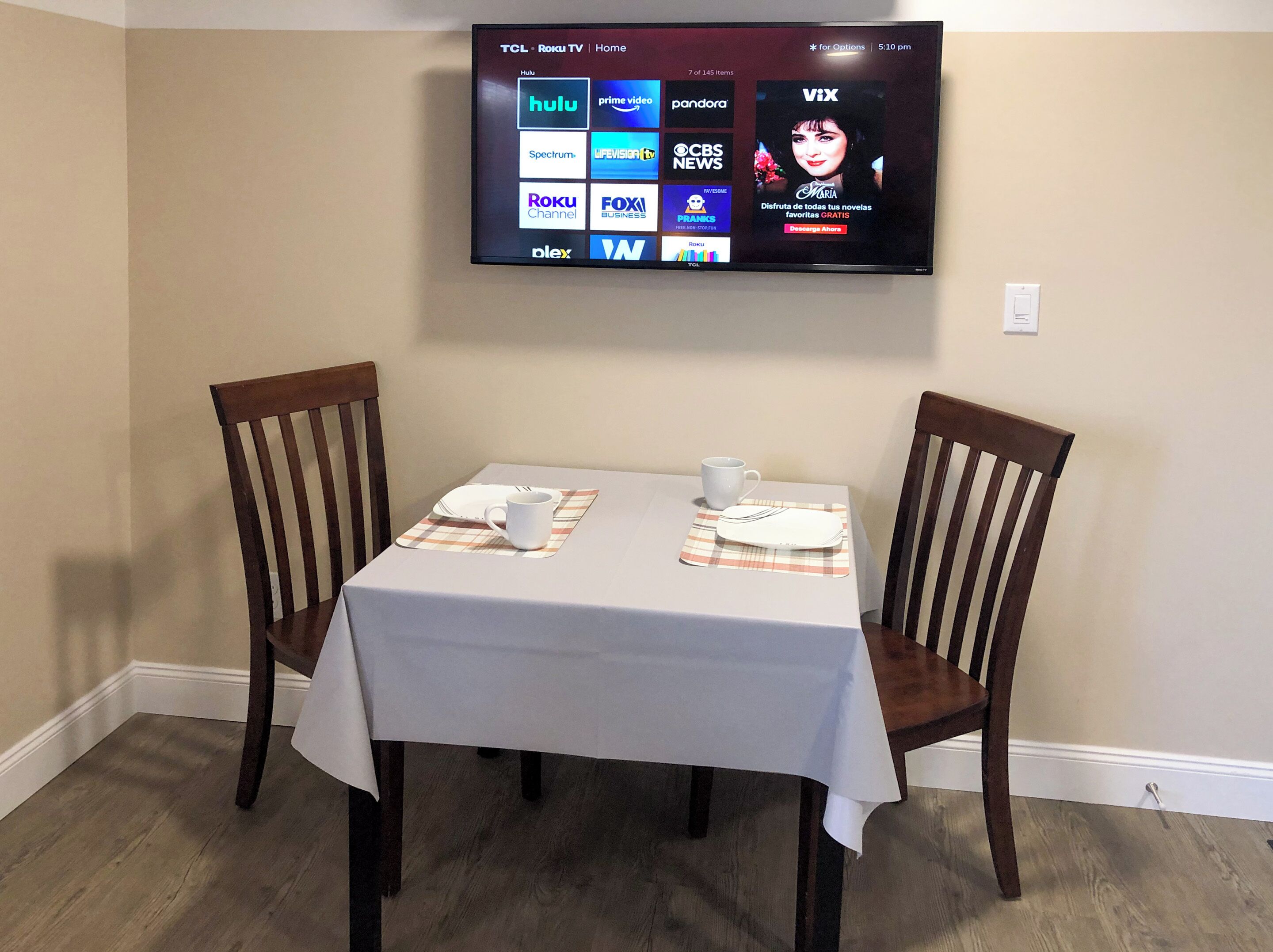 A dining area with a table set for two, a television mounted on the wall displaying various streaming services, and a pair of wooden chairs.