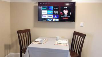 A dining area with a table set for two, a television mounted on the wall displaying various streaming services, and a pair of wooden chairs.