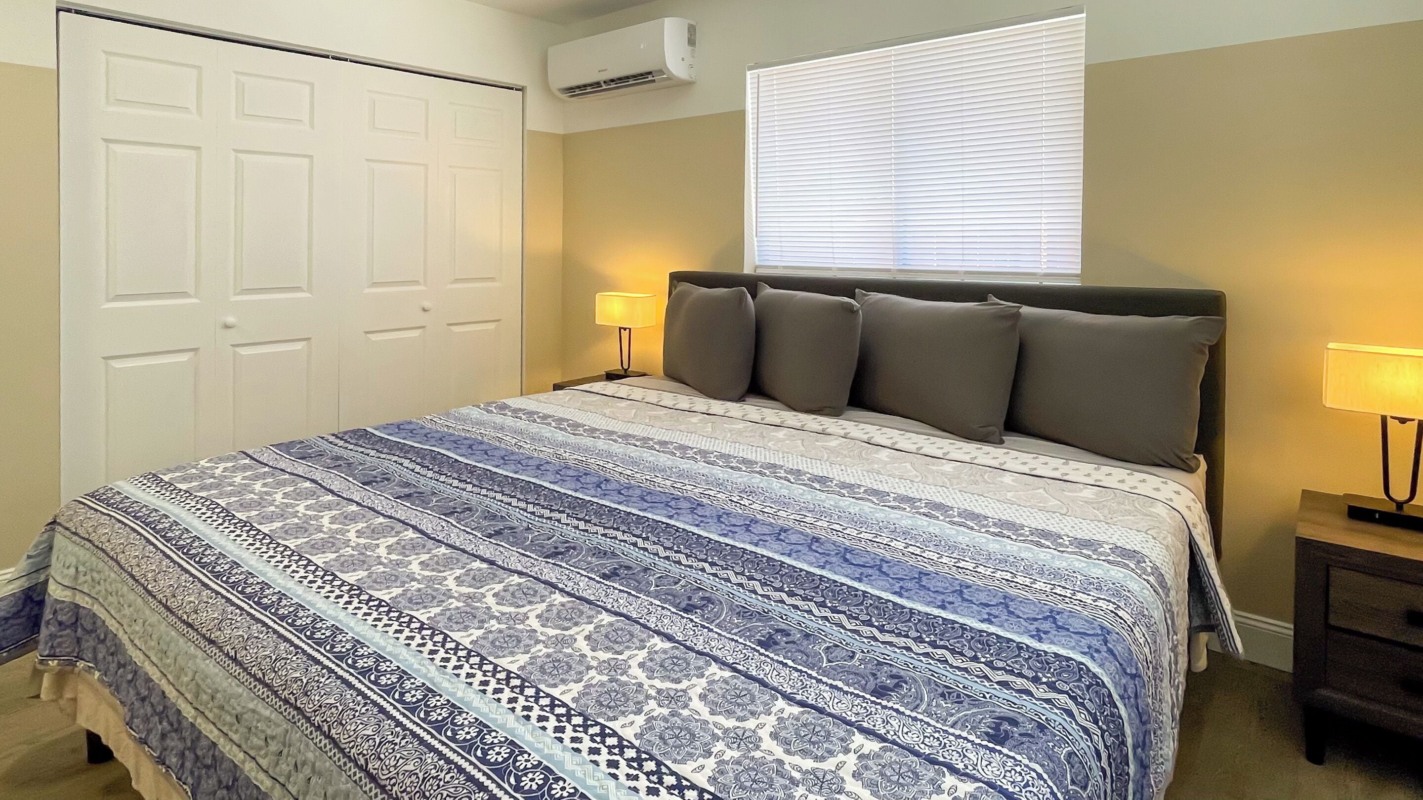 A bedroom with a patterned bedspread, a headboard with multiple pillows, a bedside lamp, and a window with blinds.