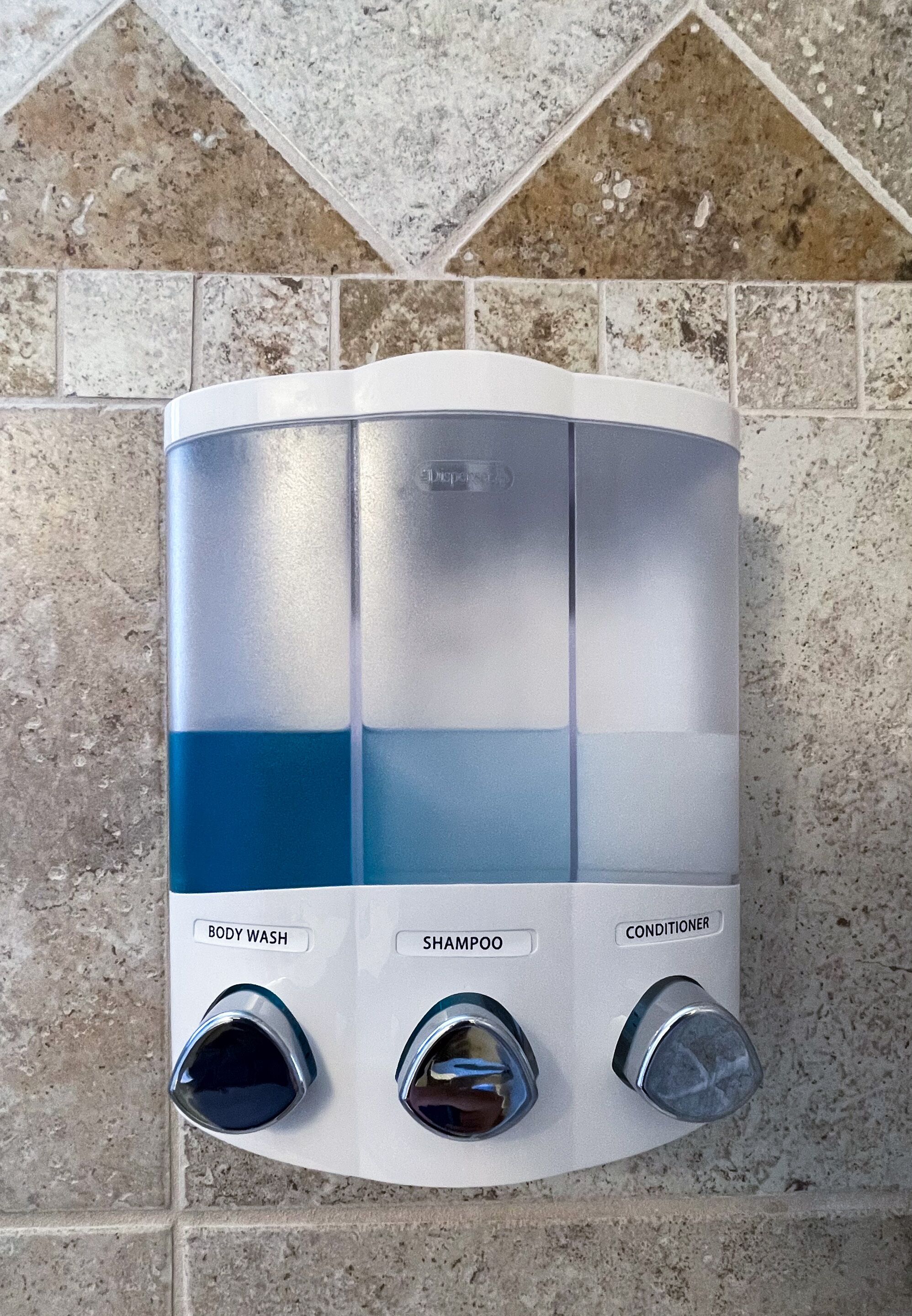 Un distributeur mural pour la salle de bain, doté de trois compartiments et de boutons étiquetés « GEL DOUCHE », « SHAMPOING » et « APRÈS-SHAMPOING ».