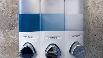 Un distributeur mural pour la salle de bain, doté de trois compartiments et de boutons étiquetés « GEL DOUCHE », « SHAMPOING » et « APRÈS-SHAMPOING ».
