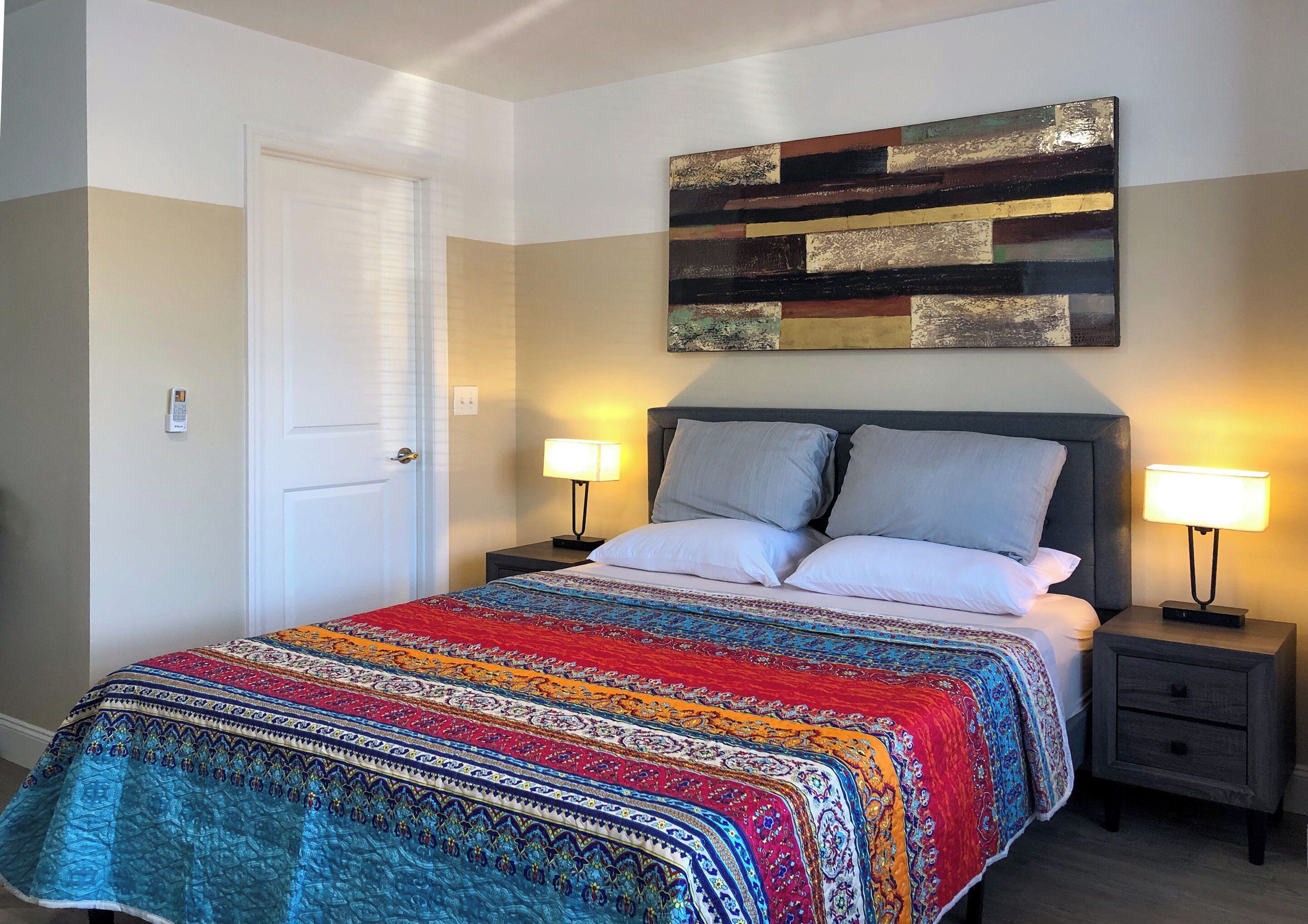 Een net opgemaakt bed met een kleurrijk dekbed, twee nachtlampjes en een modern abstract schilderij erboven.