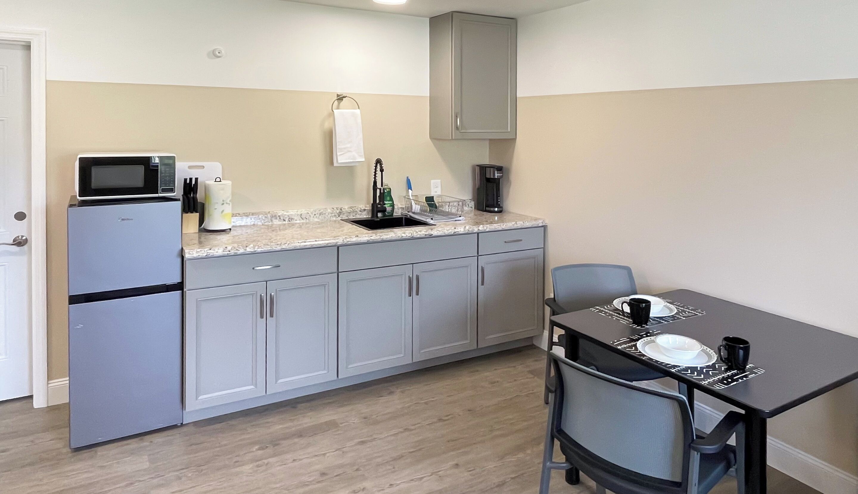 A compact kitchen with a microwave, sink, and coffee maker. There is a dining table with chairs and a small cabinet above the counter.