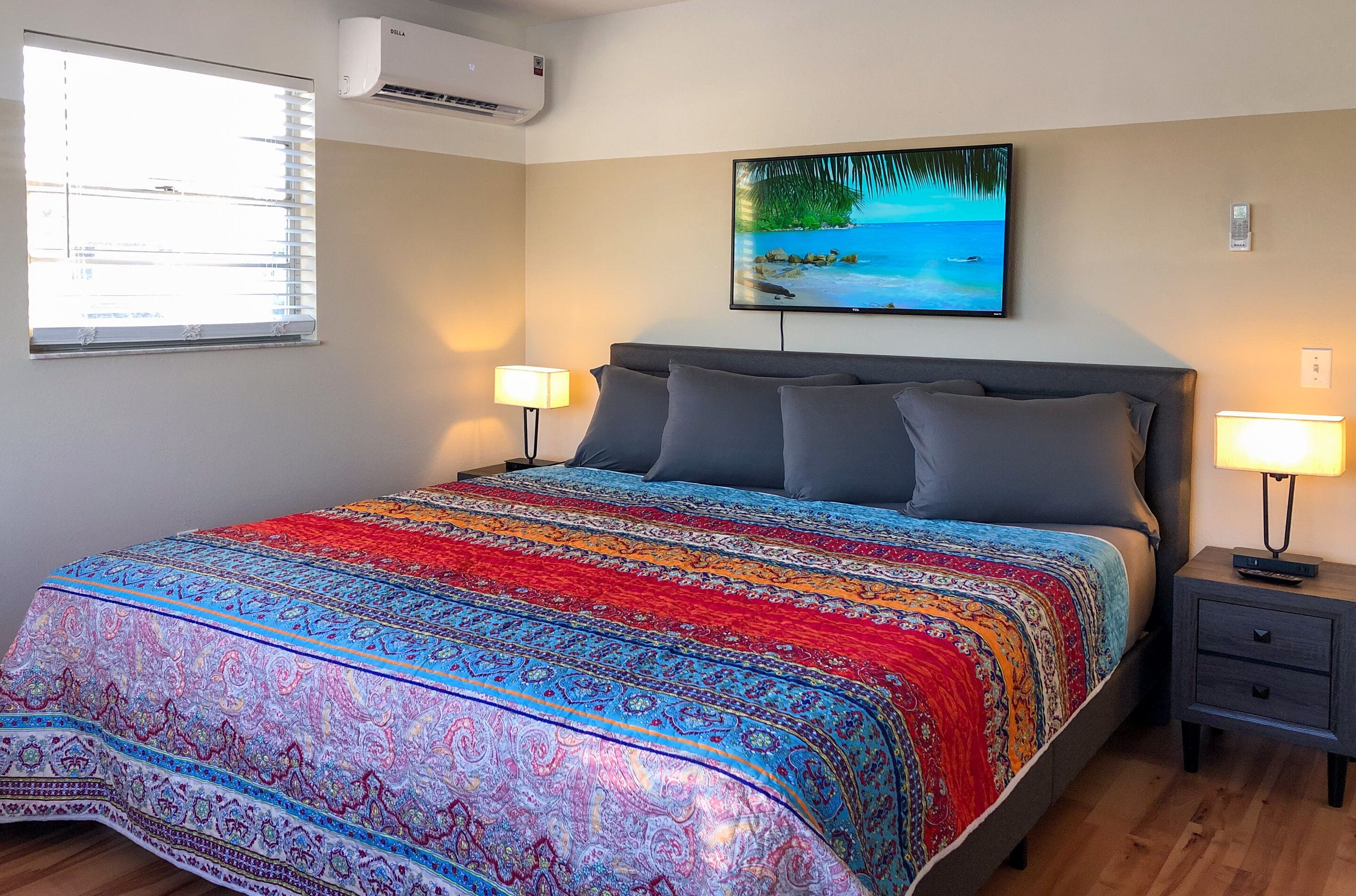 A bedroom with a large bed, two bedside tables with lamps, a wall-mounted air conditioner, and a painting of a beach scene.