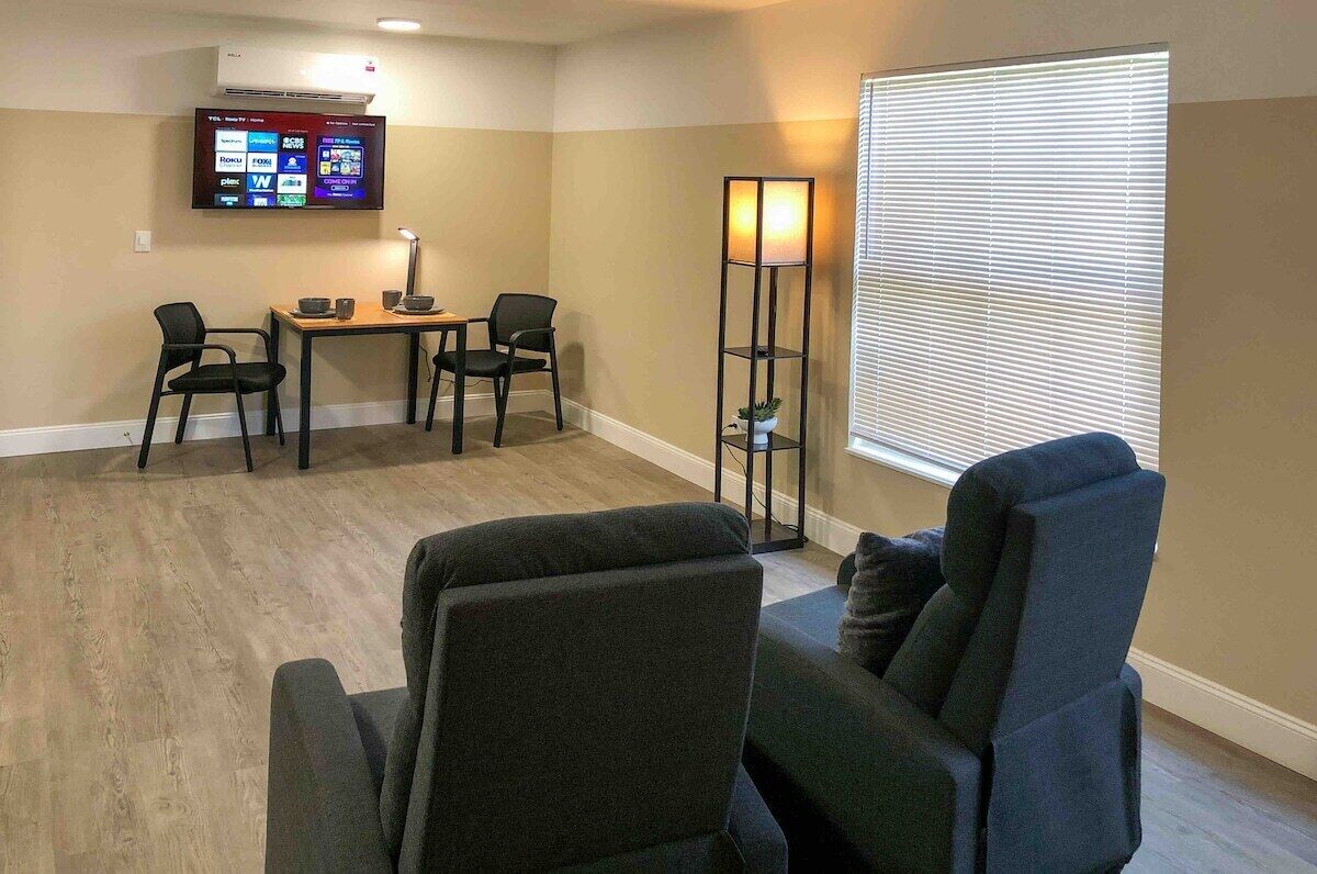 Una sala de estar con un televisor montado en la pared, una lámpara de pie, un sofá y una ventana con persianas.