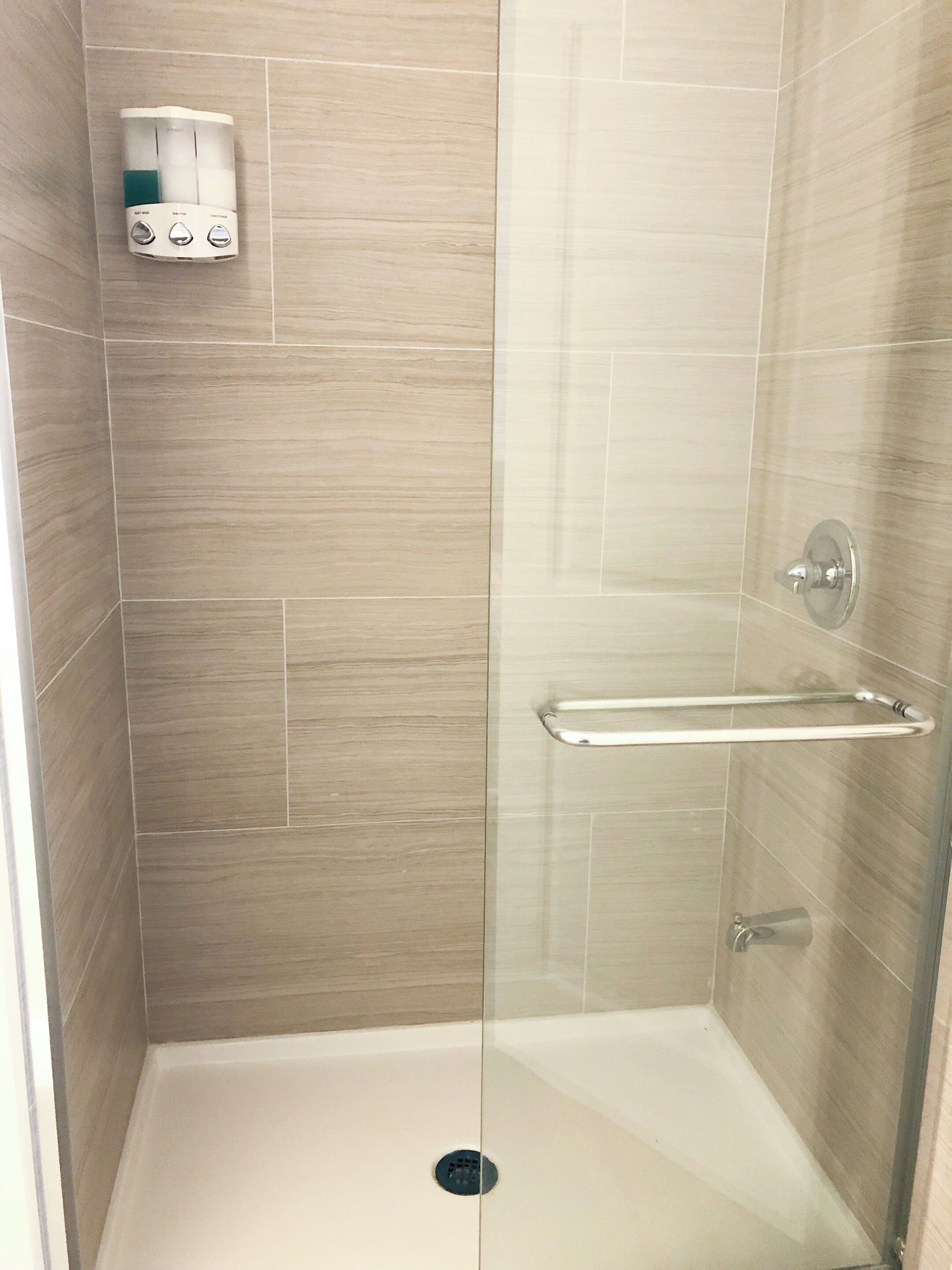 A glass-enclosed shower stall with a handheld showerhead, a towel rack, and a soap dispenser on the wall.