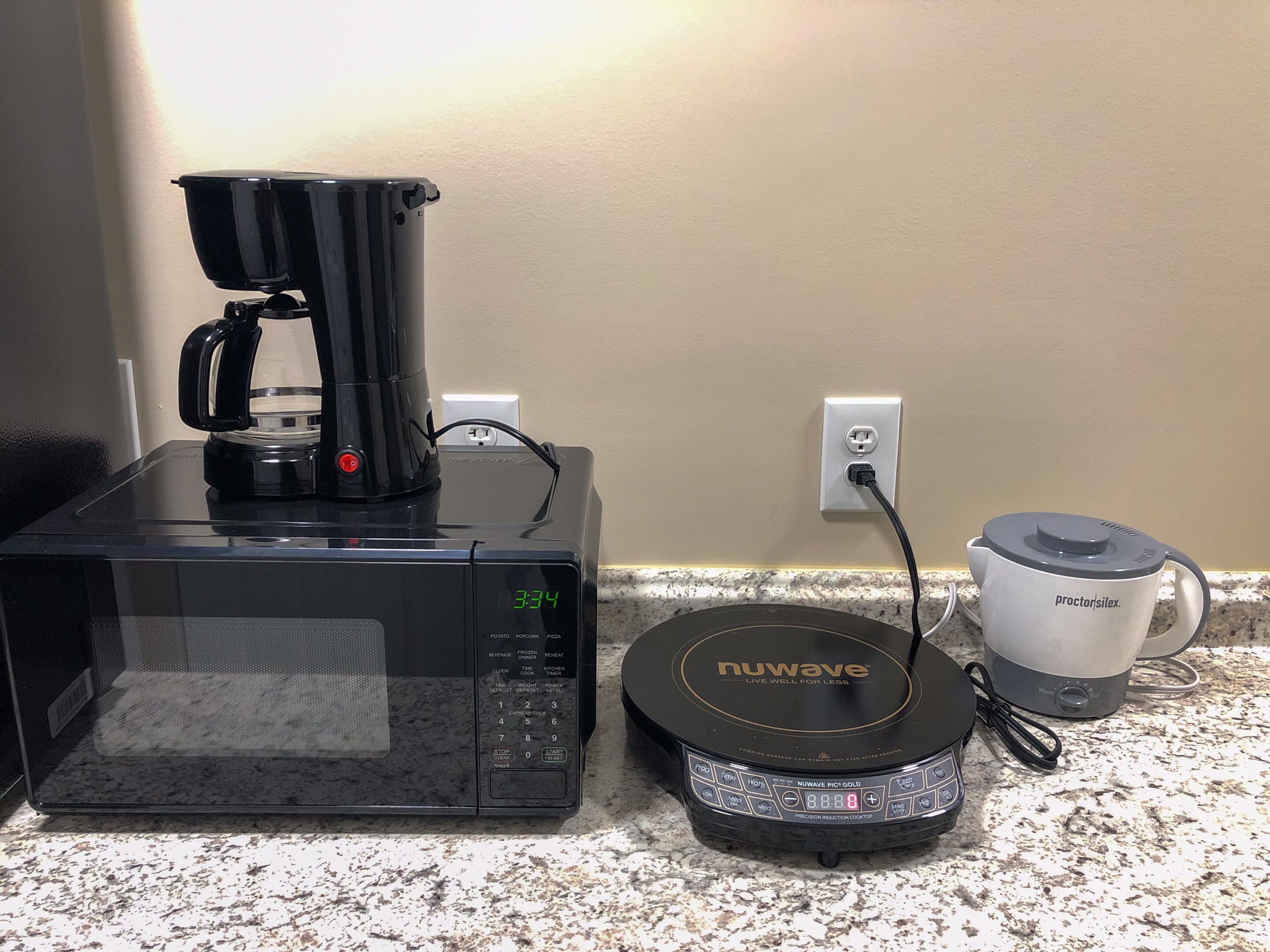 Un horno microondas con una cafetera encima, una hornalla de inducción Nuwave Precision y una taza Proctor-Silex sobre una encimera de granito.