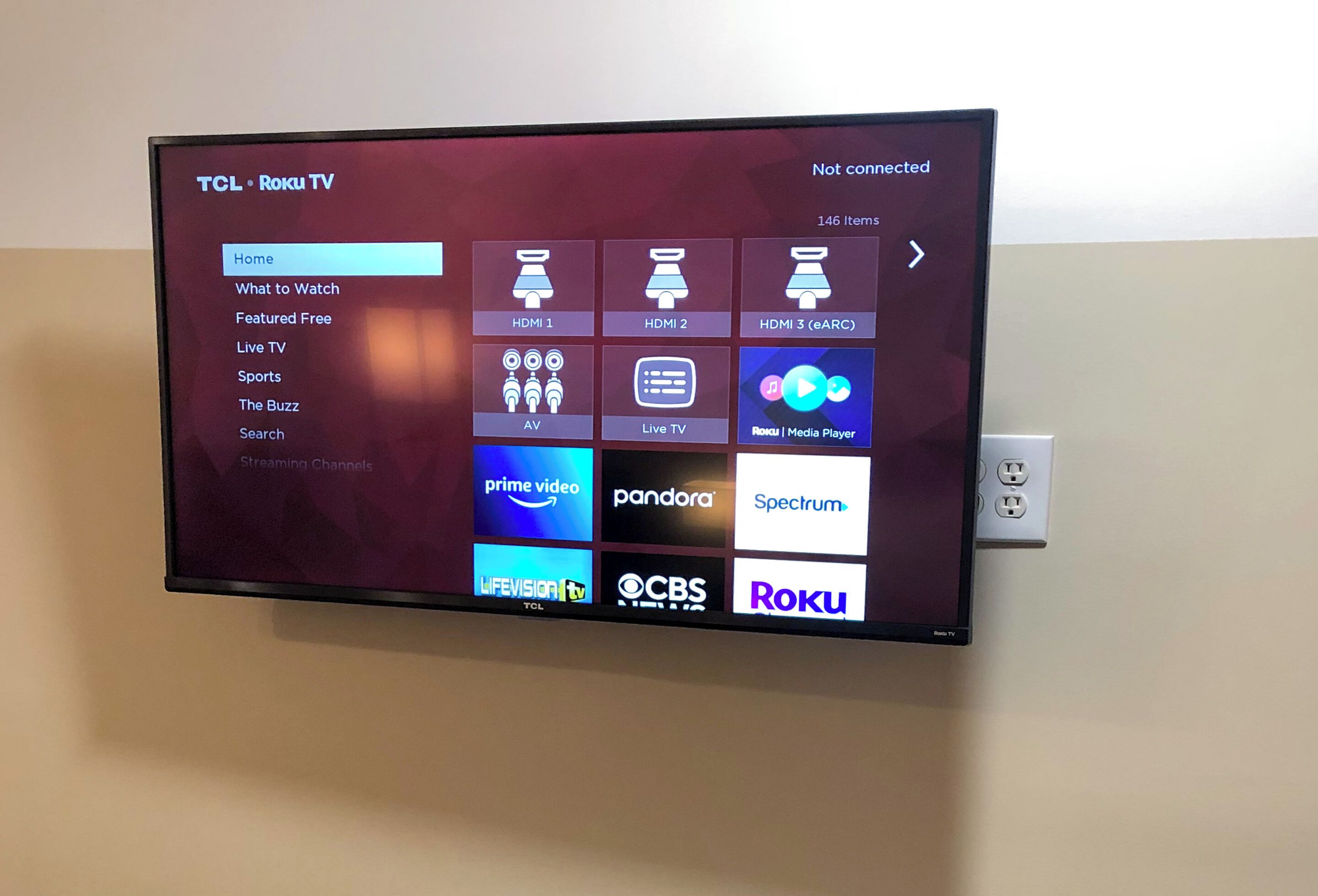A TCL Roku TV mounted on a wall with a menu interface displaying various streaming services and options.