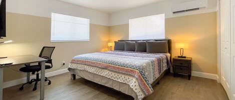 A bedroom with a bed, a desk, a chair, a nightstand, and two windows.