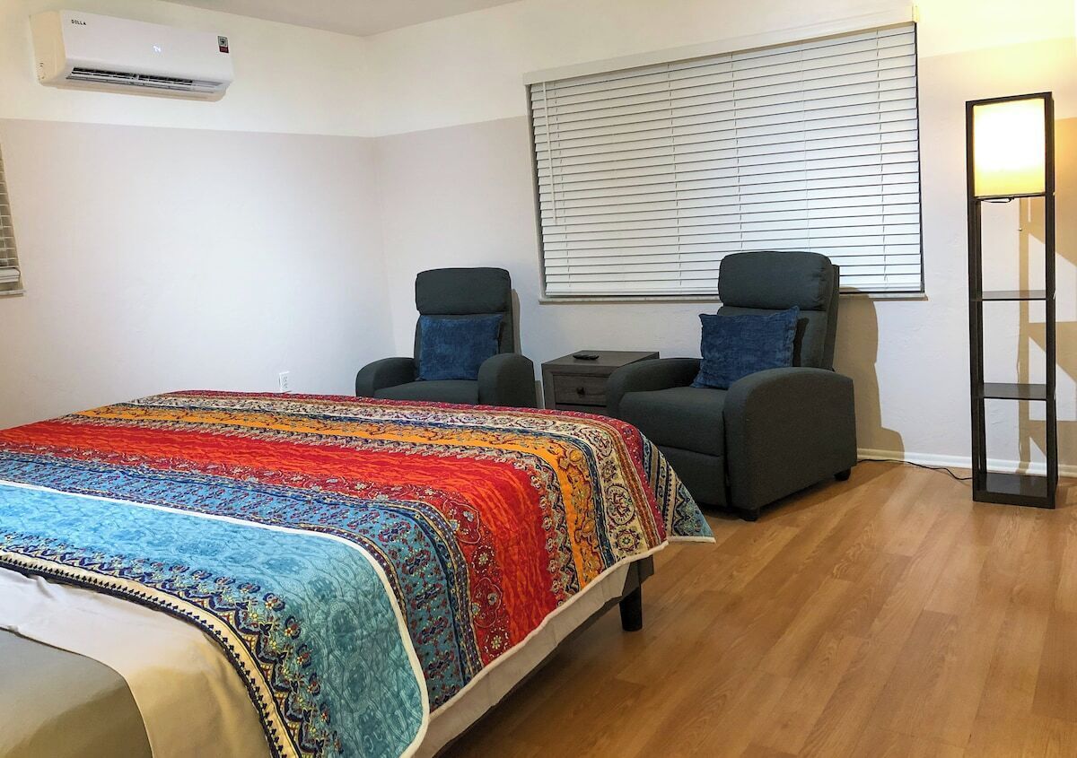Een slaapkamer met een bed, twee fauteuils, een nachtkastje, een vloerlamp en een raam met zonwering.