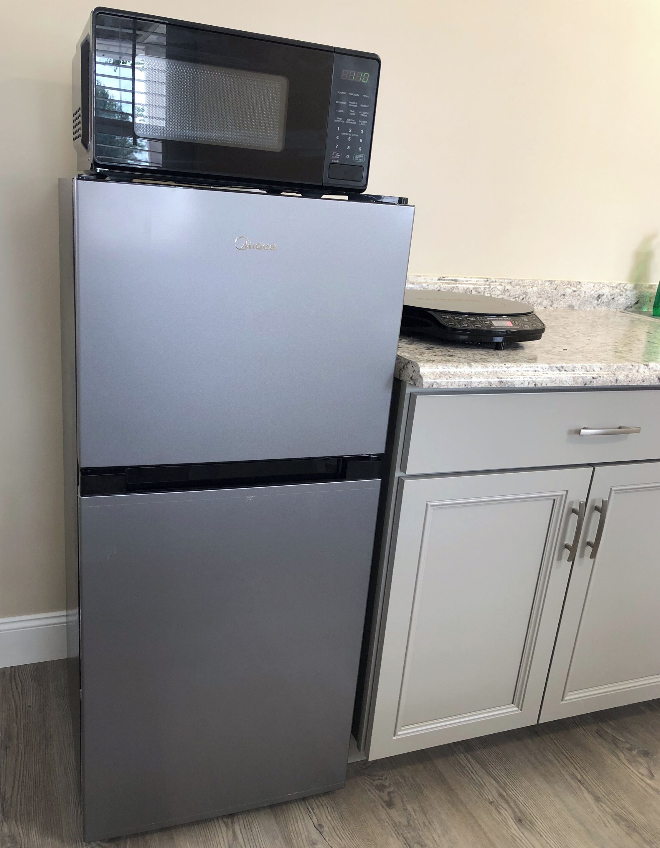 A compact kitchen with a stainless steel refrigerator, a microwave on top, and a granite countertop.