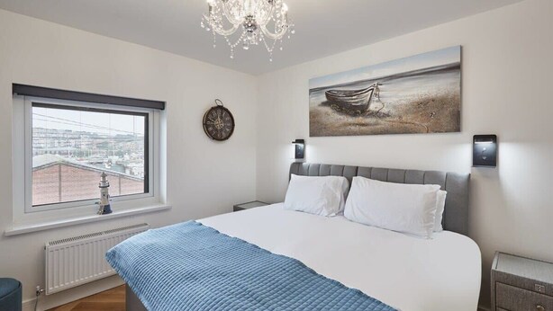 A bedroom with a large bed, a view of the city through a window, a clock, and a painting of a boat on the wall. - Host & Stay | Wadsworth Cottage (Whitby)