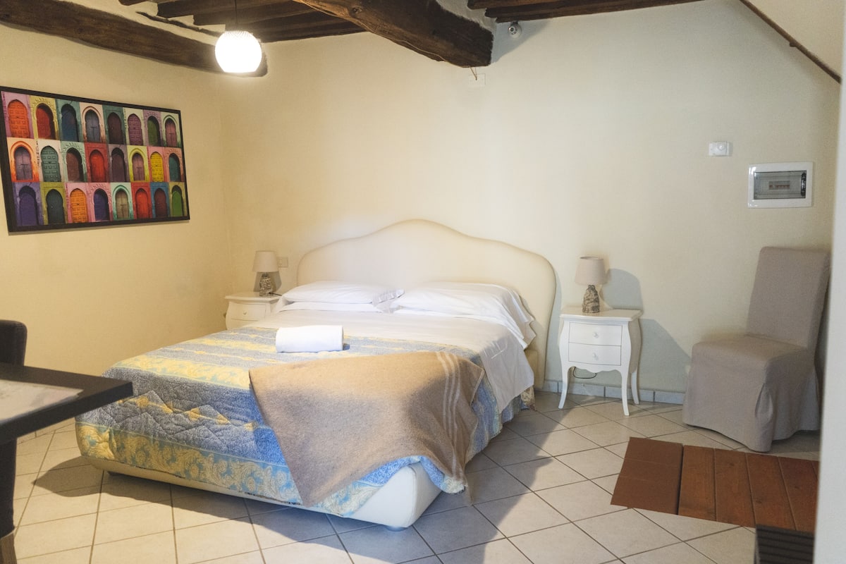 A bedroom with a bed, two bedside tables, a chair, a colorful wall art, and a ceiling light.